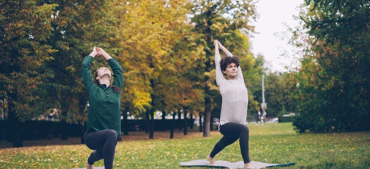 Slender girl yoga student is learning Eagle pose under guidance of experienced teacher during individual practice with instructor in park. Beautiful autumn nature is in background.