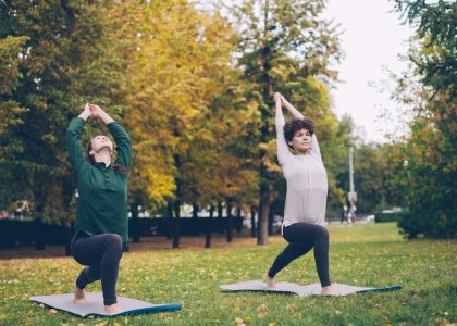 Slender girl yoga student is learning Eagle pose under guidance of experienced teacher during individual practice with instructor in park. Beautiful autumn nature is in background.